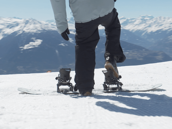 Nidecker Supermatic Snowboarder in Bindung auf einem weißen, schneebedeckten Hang mit Bergen im Hintergrund.