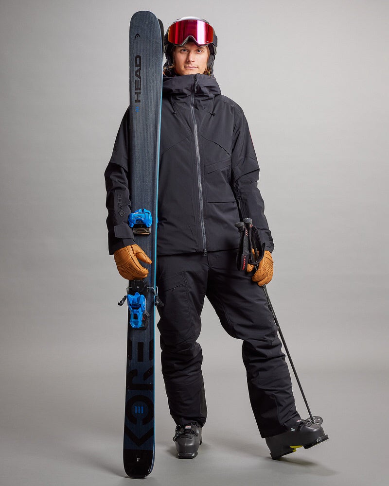 Skifahrer in Gore-Tex Person in schwarzer Gore-Tex Skihose und Jacke hält Ski und Stöcke, mit roter Skibrille und Handschuhen.