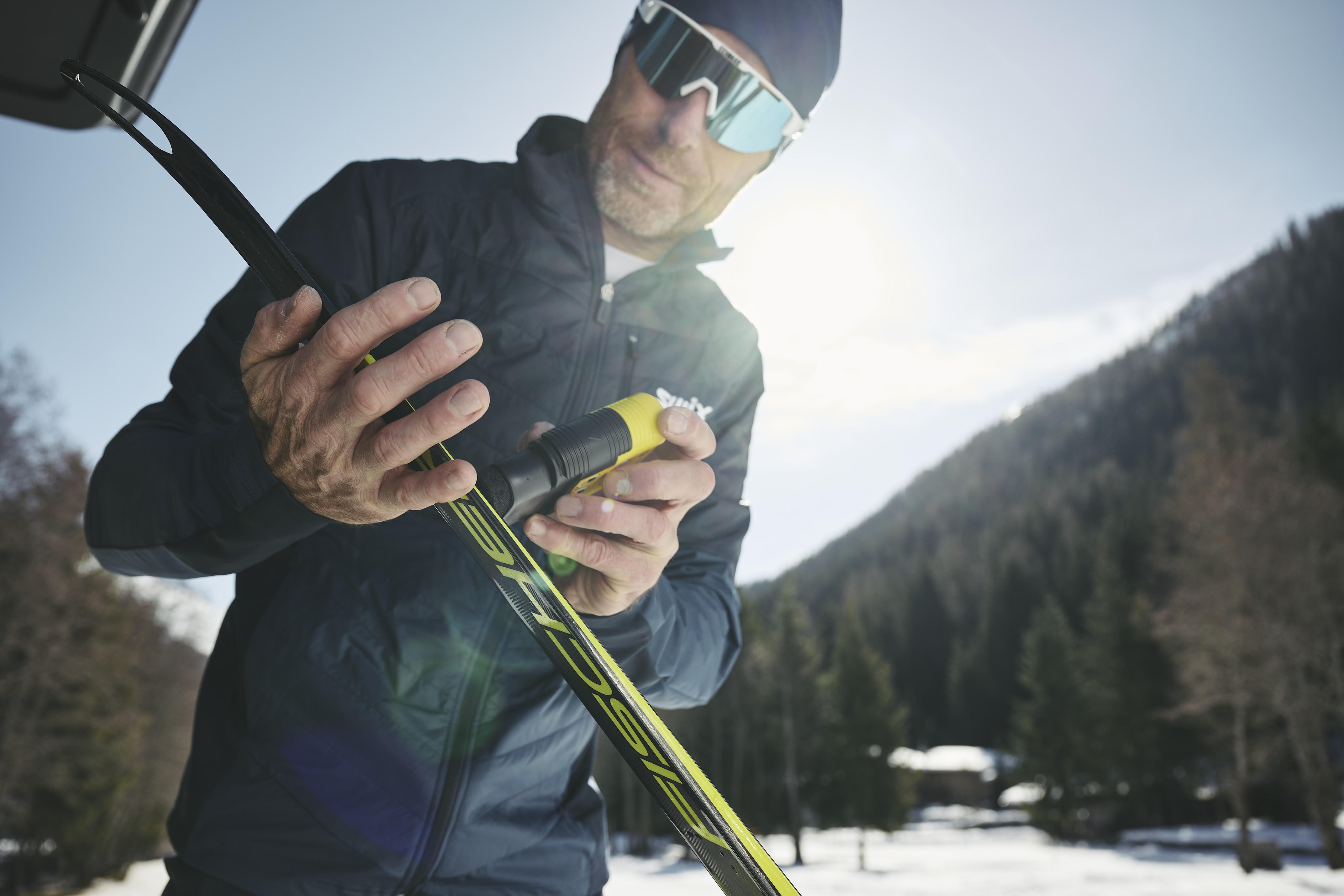 Darum Waxen mit Toko Mann mit Skibrille wachst Ski mit gelbem Wachsstift, bewaldeter Hügel im Hintergrund.