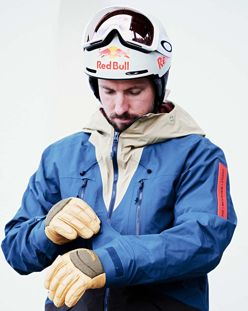 Entdecke die Marke: The Mountain Studio Ein Mann in Skikleidung, mit Helm und Brille, zieht Lederhandschuhe an. Die Jacke ist blau mit beigem Einsatz.