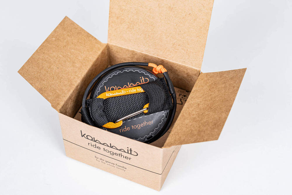 Zugsystem im Karton Geöffnete Kartonverpackung zeigt das kommit Zugsystem, ein aufgerolltes Seil mit schwarzer Schlaufe und Karabiner.