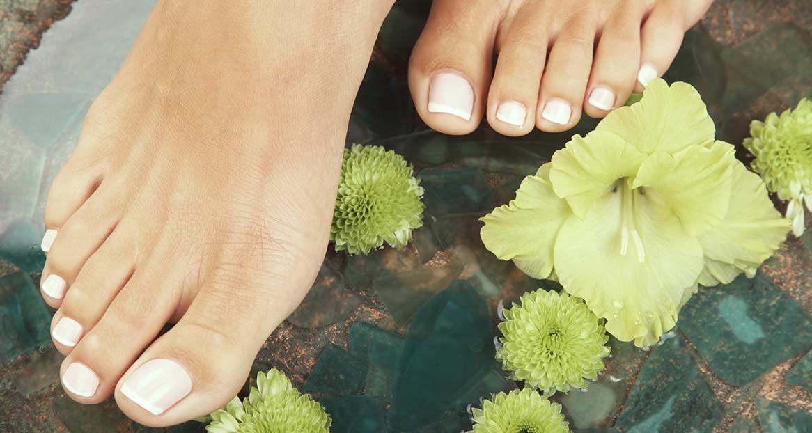 Zwei Füße mit hell lackierten Zehennägeln ruhen in Wasser mit grünen Blumen. Die Oberfläche unter dem Wasser ist mosaikartig.