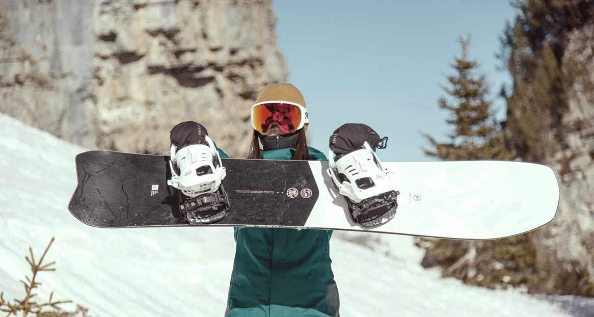 Person mit Mütze und Skibrille hält ein Snowboard mit Bindungen vor verschneiter Bergkulisse.