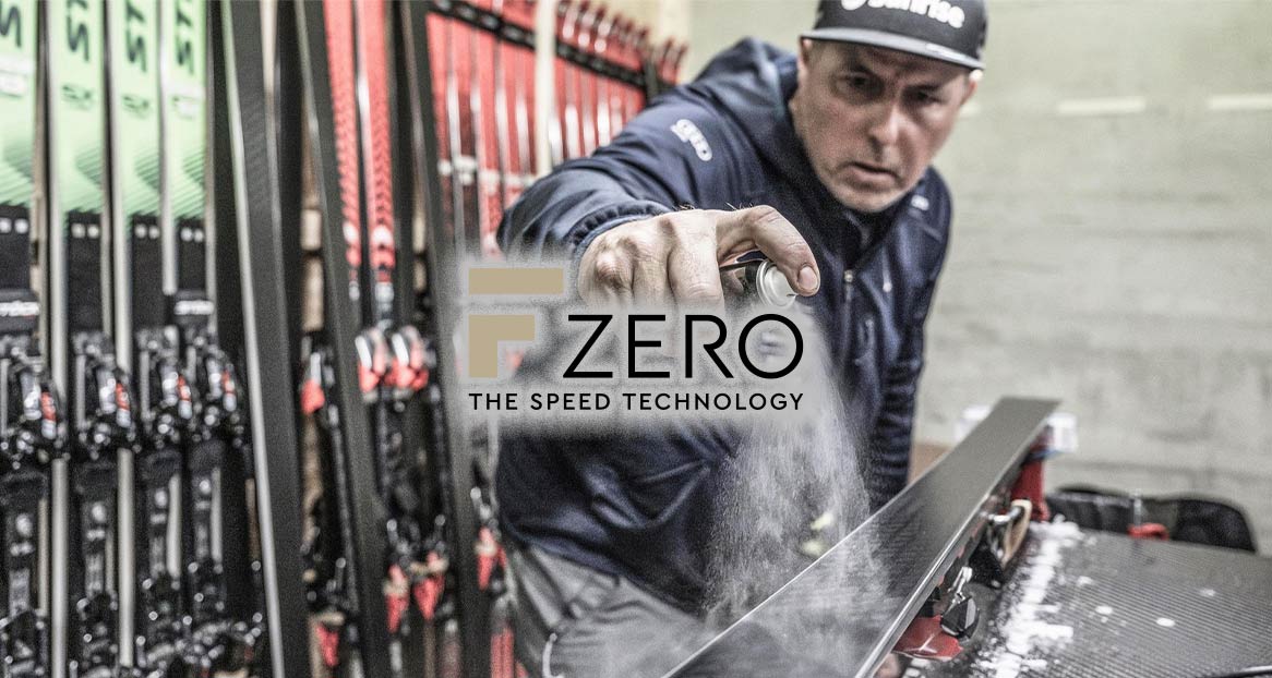 Ein Mann sprüht Wachs auf einen Ski, daneben das Logo „F Zero – The Speed Technology“ und weitere Ski.